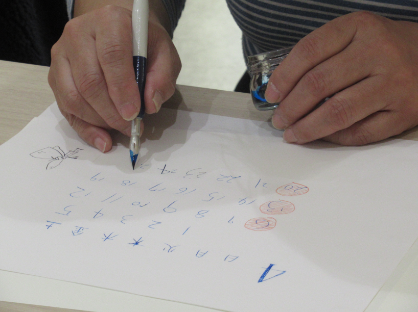 机の上で左手に青いインクの瓶を持ちながら、右手で持ったつけペンで、白い紙に4月のカレンダーの数字を青いインクで一文字ずつ丁寧に書き進めている様子を近くから捉えた写真