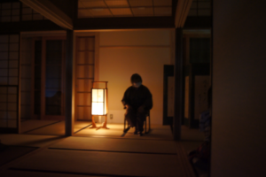 薄暗い和室で行灯の柔らかな灯りに照らされた話者の様子を正面から撮影した写真