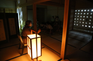 薄暗い和室で、行灯の隣に置かれた椅子に座った話者が、手振りを加えながら話をしている様子の写真