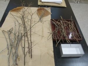 黒いテーブルの上に広げられた茶色の包装紙やトレイの上に、様々な太さや長さの乾燥した木の枝が、サイズごとに仕分けられて整然と準備されている写真