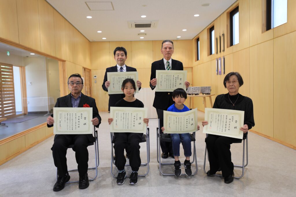 木目調の明るい室内で、表彰状を手にした老若男女6名が、前列と後列に分かれて正面を向き、晴れやかな表情の集合写真