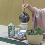 和装の女性が卓上の茶器の前で黒い茶釜を持ち上げ、丁寧に茶の準備を進める様子の写真