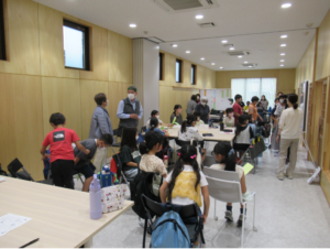 複数のテーブルと椅子が並んだ広い室内に、大勢の参加者の子どもや保護者が集まりにぎやかな様子の写真