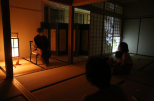 薄暗い和室で行灯の灯りに照らされた話者と参加者たちが向き合って座っている写真