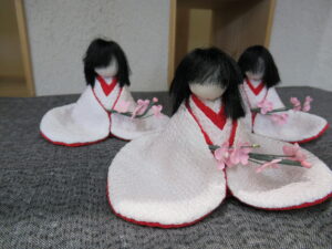 赤い襟の白い着物をまとい桜の枝を手にした三体の小さな人形が並んだ展示の写真