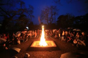 夜の広場で大きな焚き火を囲む多くの人々が傘を手に円形に集まり橙色の炎の揺らめきが深い闇の中を温かく照らす写真