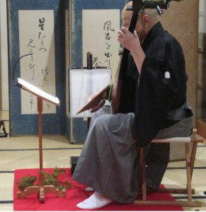 和室の畳の上に置かれた赤い毛氈に座り、紋付きの黒い和服と袴を身にまとった奏者が、目の前の譜面台に置かれた楽譜を見つめながら薩摩琵琶を静かに演奏している様子を横から捉えた写真