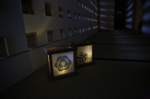 薄暗い和室の畳の上に家紋入りの二つの行灯が柔らかな光を放ち、壁に並んだ小窓からの明かりが幻想的な光模様を映し出す写真