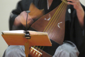 譜面台に置かれた楽譜を確認しながら、紋付き袴姿の奏者が薩摩琵琶を手に持ち、大きな撥を上から下へと素早く振り下ろして弦を弾く手元を捉えた写真