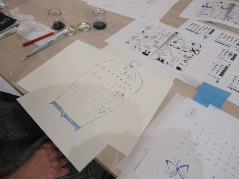 木目調の机の上に、鳥かごを模した枠の中に5月のカレンダーが手描きされた紙や、蝶のイラストが添えられた4月のカレンダーなど、複数の制作物が並び、奥にはインク瓶やペン、図案の見本が置かれている写真