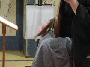 紋付き袴姿の奏者が椅子に腰掛け、譜面台の楽譜を確認しながら伝統楽器である薩摩琵琶の弦を大きな撥で弾き鳴らしている様子を横から捉えた写真