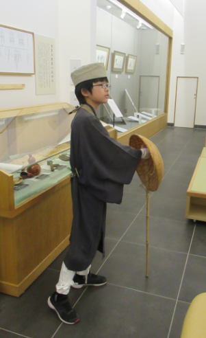 展示室で旅装束を着たこどもが、杖を手に、展示ケースを背景に立っている写真