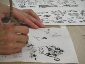 明るい色の木製テーブルの上で、手本となる花のイラストが描かれた紙を参考にしながら、参加者が細い木の枝を筆のように持って、白い和紙に墨で繊細な植物の絵を丁寧に描き写している写真