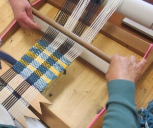 木の作業台で二人の手が卓上織機を動かし黄青白の糸が格子状に織り上がっていく様子を捉えた写真