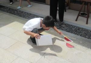 屋外の石畳に男の子がしゃがみ込み、手に持った赤いセロファン紙を貼った棒が地面に作るスイカのような形の赤い影を観察している写真