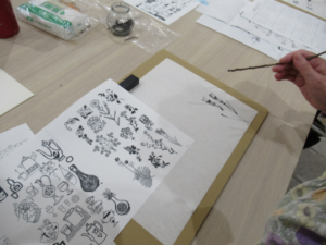 テーブルで、手本となる植物や道具のイラストが描かれた紙を横に置き、参加者が細い木の枝を筆のように持って、白い和紙に墨で草花の絵を繊細なタッチで描き写している写真