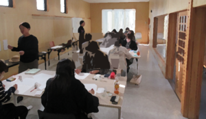 温かみがある明るい室内で、多くの人々が机に向かってつけペンの作業に集中している様子を捉えた写真