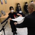 木目調の明るい室内で、黒い服に赤いリボンをつけたマスク姿の少女が、スーツ姿の男性から白い賞状を両手で受け取っている表彰式の写真