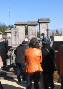 青空の下で複数の男女が古い石碑の前に並び、お参りをしている様子の写真