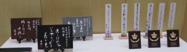 白布を掛けた展示台に、俳句が書かれた6枚の短冊、3つの王冠のエンブレムの楯、俳句が刻まれた3枚のプレートが並んでいる写真