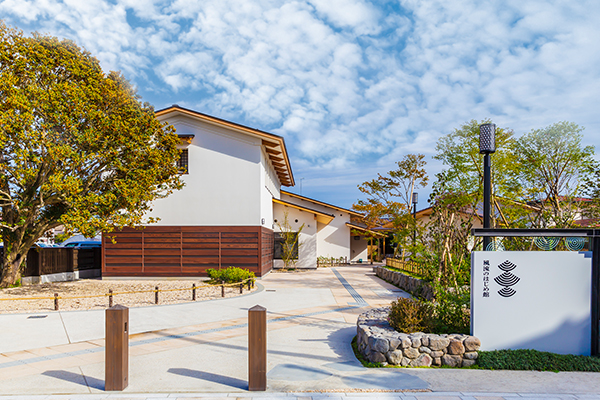 青空の下に佇む白壁と木目調の腰壁が美しい和風建築の建物で、手前には「風流のはじめ館」のロゴが入った白い看板と整えられた植栽がある施設の全景を捉えた外観写真