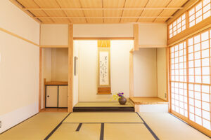 正面の床の間に一幅の掛け軸が飾られ、季節の花が活けられた花瓶が置かれている、明るく清潔感のある和室の写真