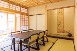 黒い長机と背もたれ付きの椅子が置かれ、床の間には一幅の掛け軸と季節の花が生けられた花瓶が飾られている、障子の後ろの窓から明るい日差しが差し込む和室の写真