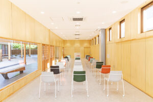 明るい木目調の壁の清潔感のある縦長の室内に、背もたれの色が異なる椅子が等間隔で整然と並べられており、左側の大きな窓からは屋外の様子が伺える郷学の間1の写真