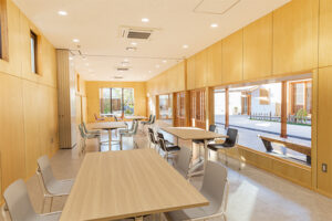 明るい木目調の壁に囲まれた開放的な室内に、複数の木製のテーブルとグレーや黒の椅子が配置され、右側の大きな窓から屋外のテラスが見える、郷学の間2の写真