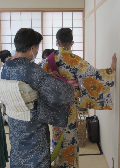 グレーの着物姿の女性が、白地にオレンジや黄色のひまわり柄の浴衣を着た女性の紫色の帯を結んでいる様子の写真