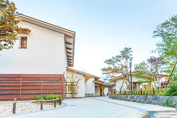 青空を背景に、白壁と木目調の腰壁が調和した蔵造り風の建物が並び、手入れされた庭木や石組みの土留めがある、「風流のはじめ館」の敷地内を捉えた外観写真