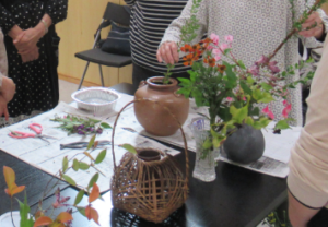 新聞紙を敷いたテーブルの上に、茶色の壺形や籠が付いたもの、細長いガラス製など様々な花瓶やハサミが置かれ、テーブルを囲んだ参加者たちに、講師の女性が花を生けながら説明をしている様子の写真