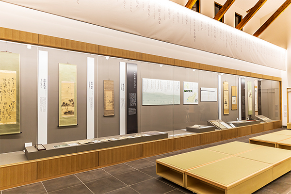 木の梁が美しく露出した高い天井のモダンな和風展示室で、壁沿いの長いガラスケース内には掛け軸や古文書が整然と並び、中央通路の畳敷きベンチと柔らかな照明が落ち着いた和の空間を演出している、資料館の内観写真