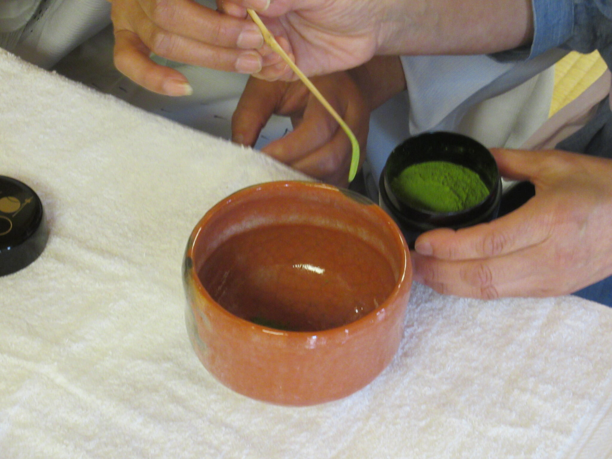 茶碗の中に緑色の抹茶を入れ、竹製の茶杓で点てる準備をする手元と、抹茶が入った黒い容器に手を添える様子を捉えた写真