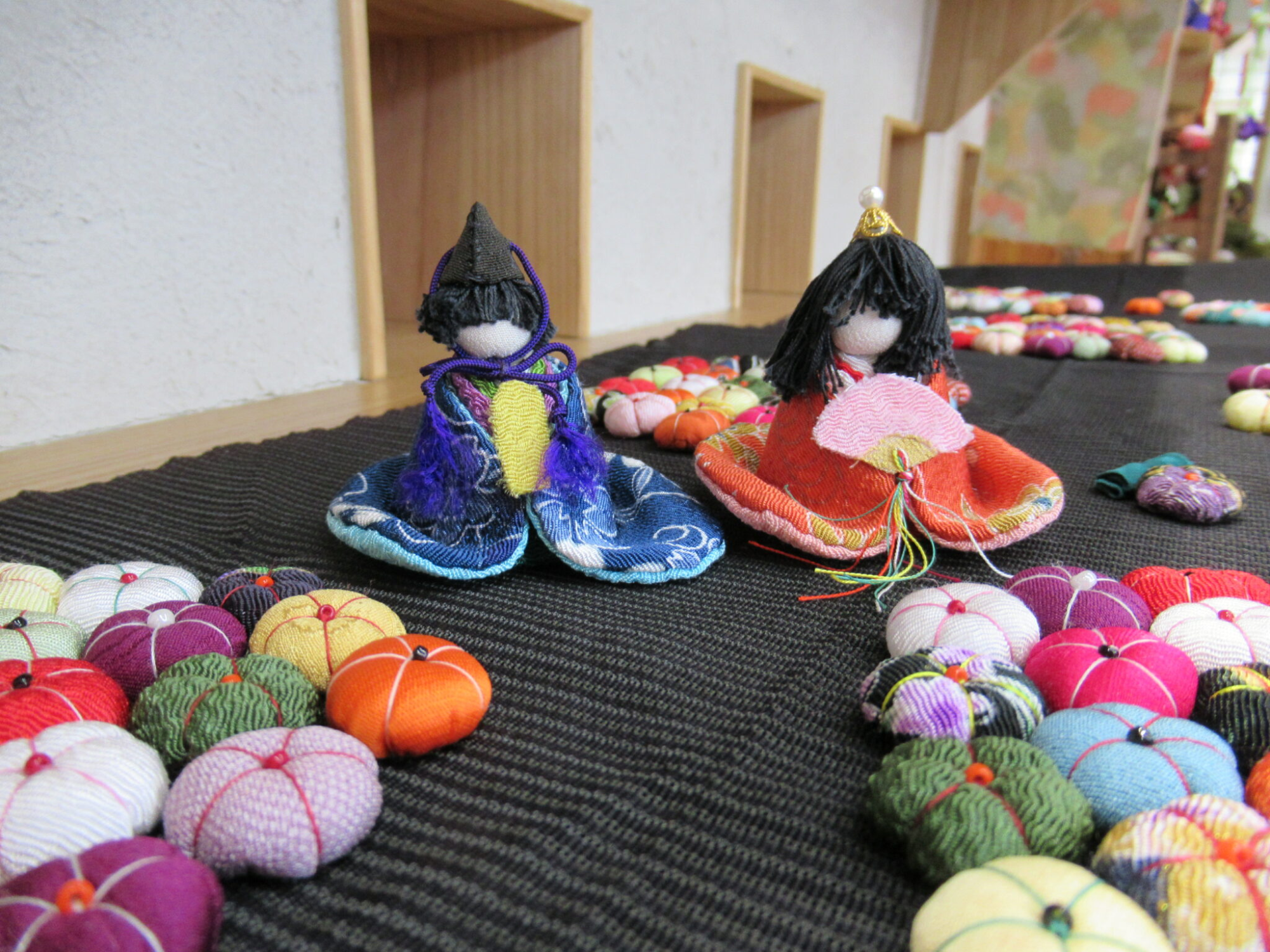 色鮮やかなちりめんで作られた男女一対の小さなひな人形が、黒い布の上に、たくさんのカラフルなちりめんの花と一緒に置かれた展示の写真