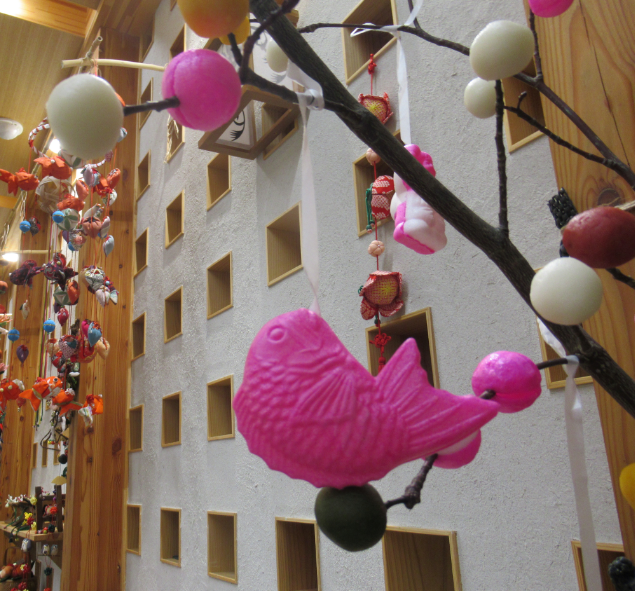 小さな四角い穴が並んだ壁の前に、黄色や白、ピンクのだんごが刺さった木の枝から鮮やかなピンク色の鯛の形をした飾りが吊るされた、だんごさしが飾られている写真