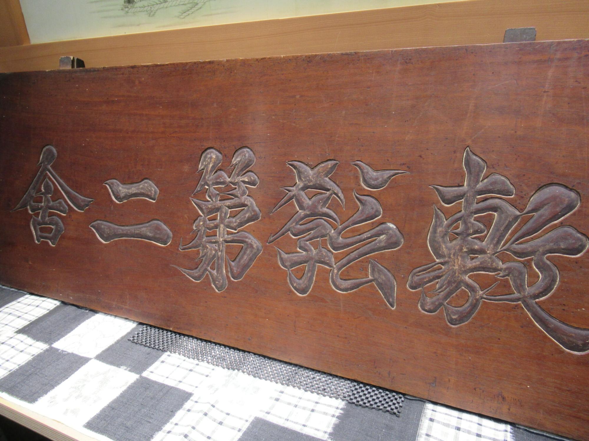 歴史を感じさせる重厚な茶色の木板に「敷教第二舎」という文字が右から左へ一文字ずつ力強く彫り込まれている看板を斜めから捉えた写真