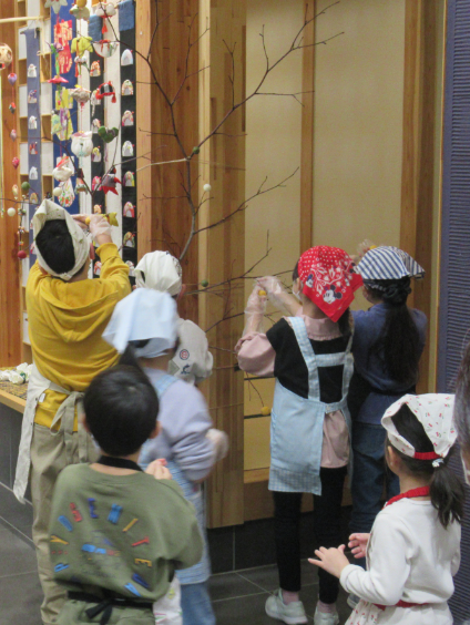 エプロンと三角巾を身に着けた子どもたちが、室内の柱に立てかけた木の枝に、色とりどりのだんご飾りを取り付けている様子の後ろ姿の写真