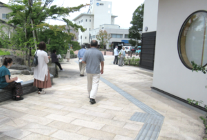 建物横の石畳の歩道を歩く人や左側のベンチに座る人の後ろ姿の写真