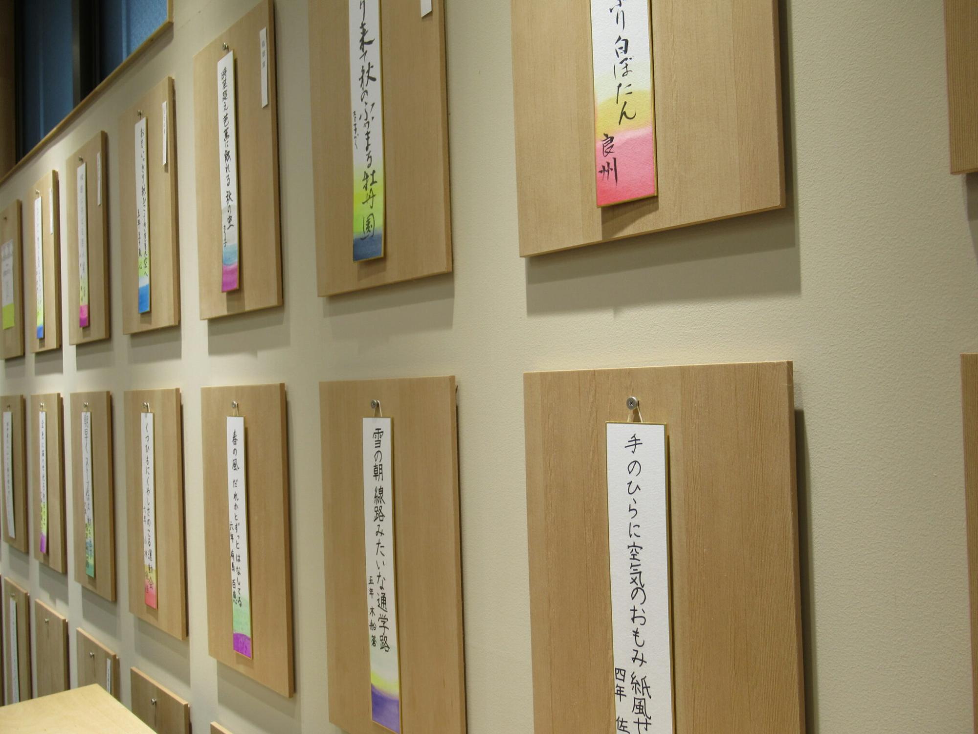 明るいベージュの壁面に、俳句が書かれた色鮮やかな短冊が貼り付けられた木製の板が、上下二段に整然と並んで展示されている写真