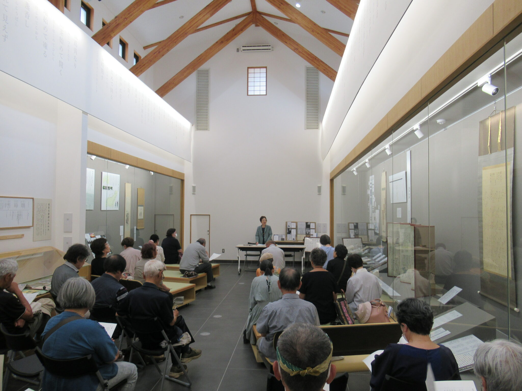高い天井に木の梁が見える、白い壁の明るい展示室で、たくさんの参加者が椅子やベンチに座り、前方に立った講師の鈴木邦子さんの話を聞いている様子の写真