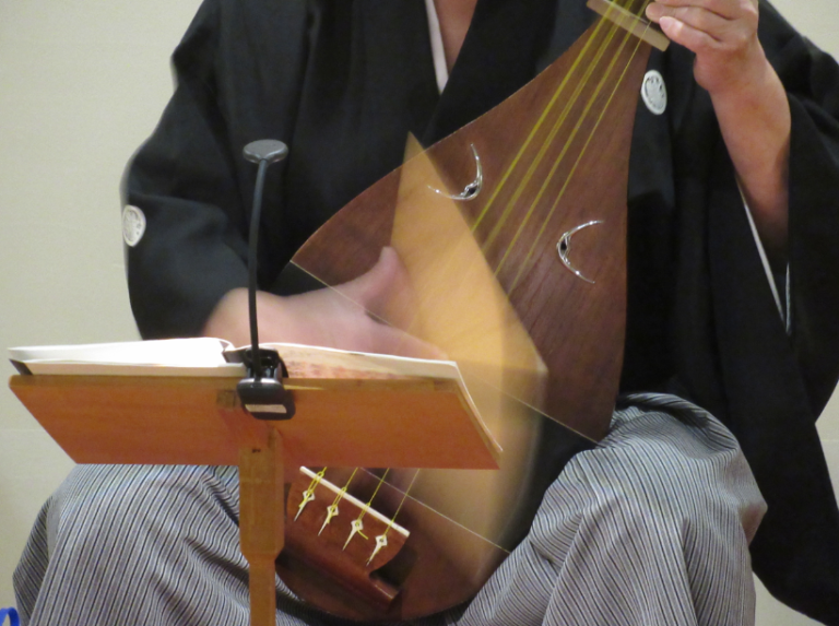 紋付きの黒い和服と縞模様の袴を着用した人物が、手元の譜面台に置かれた楽譜を確認しながら、木目が美しい撥を素早く動かして伝統的な弦楽器である琵琶を力強く演奏している、その躍動的な手の動きを捉えた写真