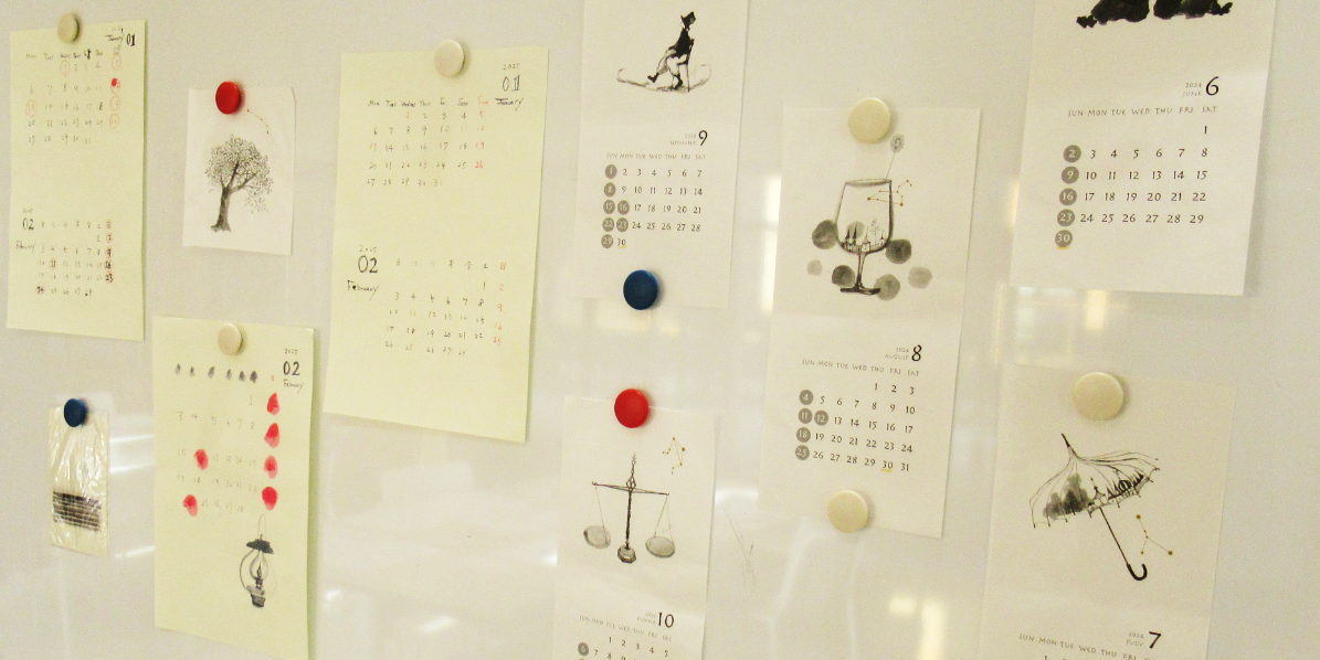 ホワイトボードに、傘や天秤、グラス、木、などの繊細なタッチで描かれたイラスト付きの様々な月のカレンダーが、カラフルなマグネットでランダムに多数貼り付けられている写真