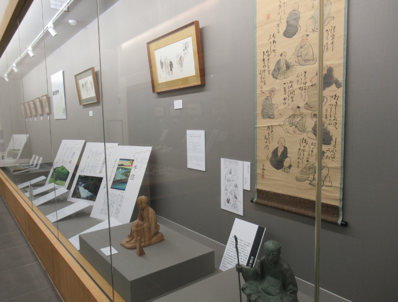 展示ケース内に掛軸や絵画、解説パネル、木彫の像などが整然と配置されている展示の様子の写真