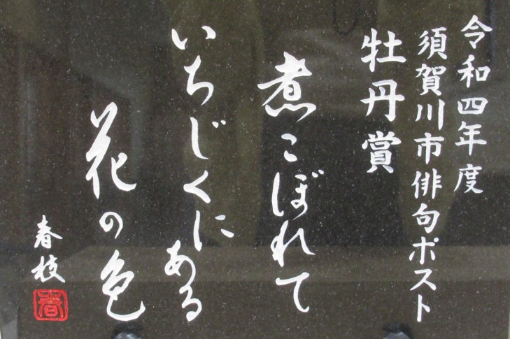 左側に「令和四年度須賀川市俳句ポスト牡丹賞」と記され、中央に「煮こぼれていちじくにある花の色春枝」と白文字で刻まれた、光沢のある黒い石板の写真