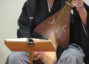 紋付き袴姿の奏者が、譜面台の楽譜に目を向けながら大きな撥を素早く振り下ろし、木目の美しい薩摩琵琶の弦を力強く弾いている写真