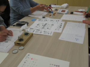 明るいベージュの机を囲んで複数の参加者が座り、それぞれつけペンを使ってカレンダー制作に熱心に取り組んでいる写真