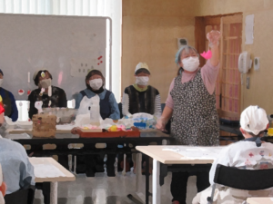 エプロンとマスクを着用した講師の女性が、ピンク色の鮮やかな鯛の飾りを高く掲げて、周りに座った参加者に示している様子の写真