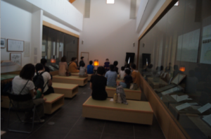 灯籠の明かりで薄暗い展示室の長椅子に来場者が整然と座り、前方の話者の話に耳を傾けている様子の写真