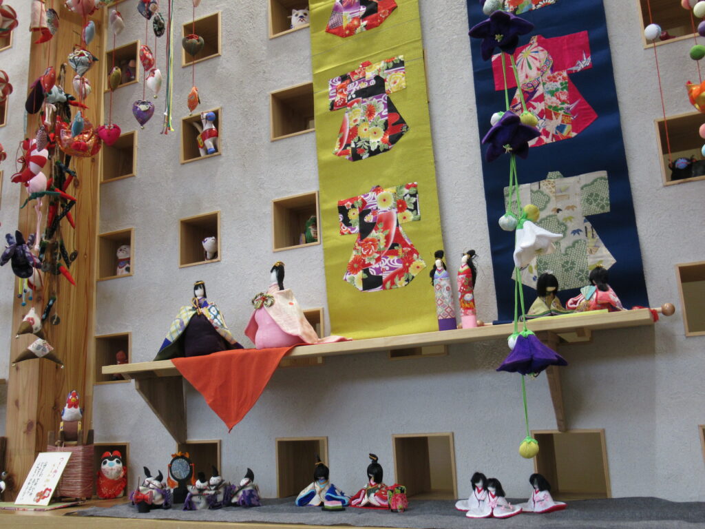 壁面の小窓に小さな飾りが並び、色鮮やかな着物の人形や、つるし飾りがいくつも飾られた華やかな展示の写真